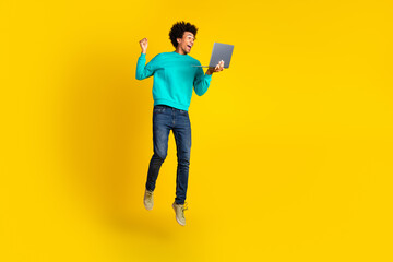 Happy young man with laptop in teal sweatshirt on vibrant yellow background jumping with joy