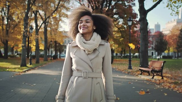 Bellissima donna di origini Afro americane vestita con un elegante cappotto e una sciarpa cammina felice in un parco di una citt&agrave; in autunno durante il foliage