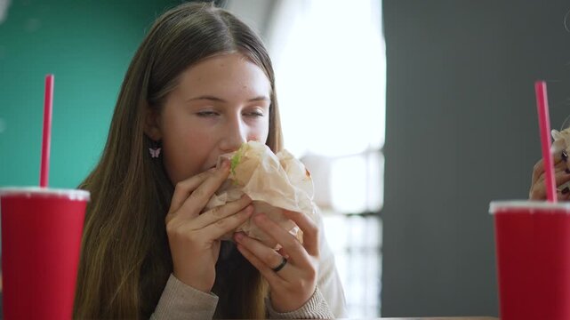 Eating burger with smiling teenager girl holding wrapped burger and red drink on table in fast food restaurant at lunch while enjoying snack and glance with relaxed expression and casual clothing