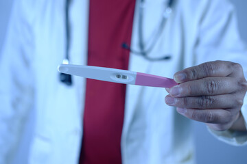 A male doctor holds a pink pregnancy test and gestures to his patient indicating a positive result....