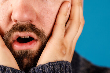 A close-up of a man's face showing shock, holding his face with hands, against a blue backdrop....
