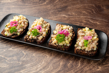Appetizer of rye bread topped with chopped pickles, onions, fish, parsley, and pickled red onions on a black plate. Traditional gourmet snack, neatly plated for food photography.