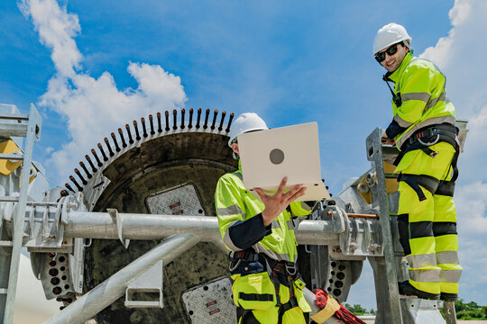 Two engineers work on a wind turbine component, with one using a laptop and the other inspecting parts on a ladder. renewable energy technology, wind power projects, and teamwork in the energy sector.