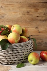 Fresh ripe apples with green leaves in wicker basket on white wooden table, closeup