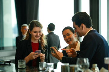 Group of colleagues sharing a laugh and using mobile phones to pay bills at stylish rooftop lounge, representing digital lifestyle, teamwork, and business socializing.