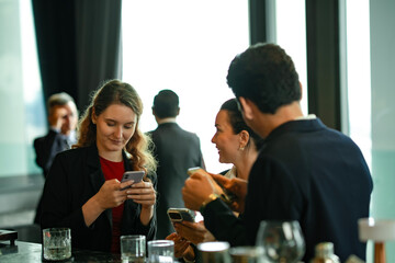 Business professionals enjoying after-work drinks at rooftop bar, smiling while using smartphones for mobile payment and digital networking in a modern urban lifestyle setting.