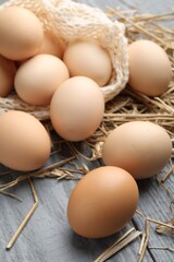 Raw chicken eggs, mesh bag and straw on grey wooden table, closeup