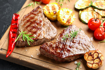 Delicious grilled beef steaks with herbs and vegetables on dark textured table, closeup