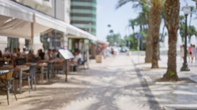 Blurred scene of outdoor cafe with people sitting and walking along palm-lined street on a sunny day with defocused background.