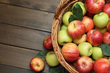 Fresh apples in wicker basket and green leaves on wooden table, top view. Space for text