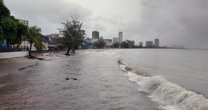 Churning grey waves encroach on an urban shoreline under a cloudy sky, with the high-rises of a modern city veiled in the distant haze. ( in the slow motion)
