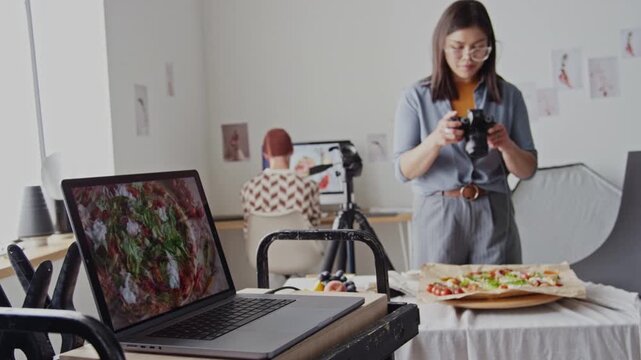 Brunette professional photographer standing near table with pizza on baking parchment and holding camera above it to take perfect shot while her colleague sorting photos on computer in background