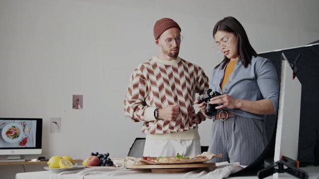 Two adult food stylist and photographer standing near table with edible props and having conversation, discussing best camera angles and adjusting decoration on table