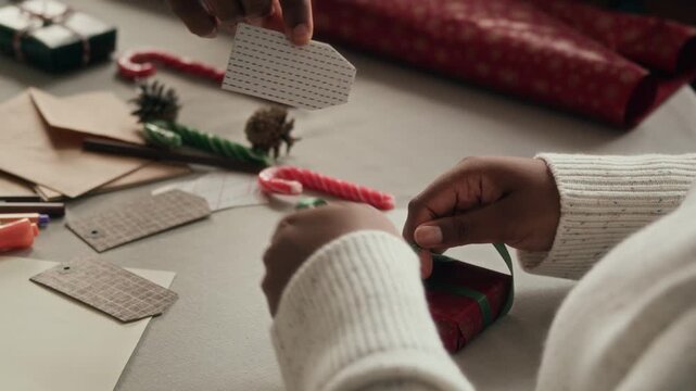 Hands of Black woman tying green ribbon on gift box decorating Christmas presents together with family sitting at table cluttered with cards and envelopes