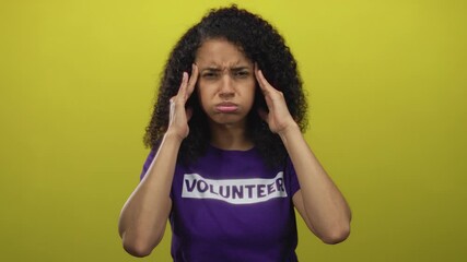 Woman wearing volunteer shirt stands against vibrant yellow background, appearing stressed, showcasing emotion with hands on temples.