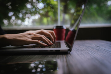 Close-up of a person typing on a laptop keyboard while holding a red coffee cup, working in a cozy...