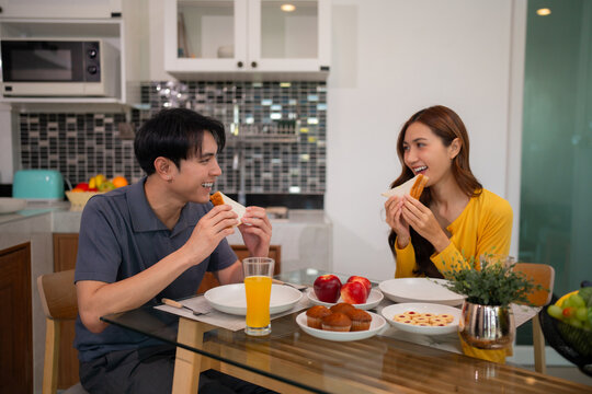 Asian adult couple eating breakfast sandwiches muffins juice happy smiling sharing morning meal home kitchen dining table enjoying domestic lifestyle healthy food - Powered by Adobe