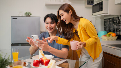 Asian adult couple enjoy morning breakfast together at home sharing online content on smartphone creating happy relaxing domestic lifestyle moments