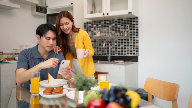 Asian adult couple enjoy morning breakfast using smartphone together at modern home kitchen relaxing domestic life - Powered by Adobe