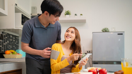 Asian adult couple share happy breakfast moment kitchen modern home using smartphone digital communication lifestyle sweet connection togetherness