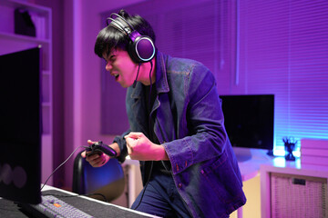 Happy asia guy gamer wear headphone participation play video game colorful neon lights computer in living room at night modern house. Esport streaming game online, Home quarantine activity concept.