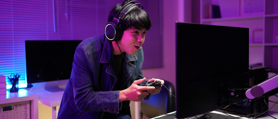 Attractive Asian male gamer enjoys playing online games on his computer feeling happy while live...