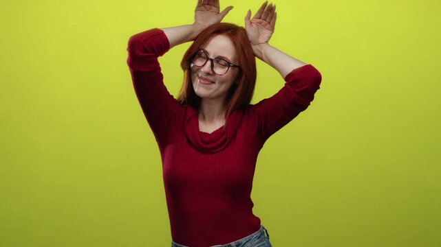 Woman smiling wearing glasses and red sweater playfully placing hands on head against yellow background showcasing her red hair.
