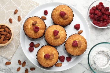 Fresh muffins with raspberries, almonds and flour on white marble table, flat lay