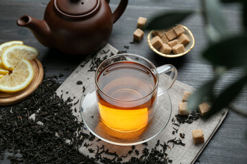 Aromatic black tea in glass cup, dried leaves, brown sugar, lemon and teapot on wooden table, closeup