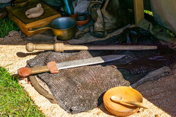 Roman gladius swords and chainmail lying on mat beside camp utensils and accessories during outdoor historical reenactment.