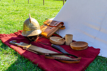 Set of historical Roman soldier items by the tent — helmet, belt, sword, horn, leather bag and wooden bowls on a red blanket.