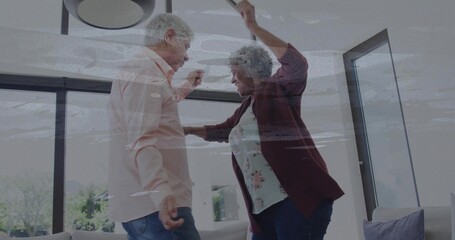 Dancing senior couple moving in living room, with large windows, sofa, pillows and ceiling light