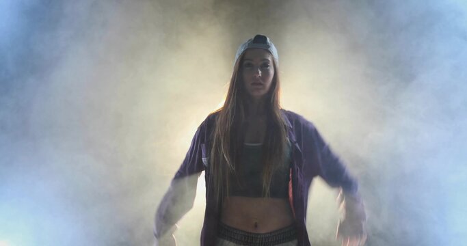 Performing female dancer wearing backwards cap and zip-up jacket, holding arms out on foggy stage - Powered by Adobe