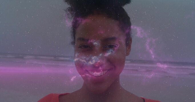 Smiling woman wearing red top standing on sandy beach edge with waves, pink-purple nebula overlay - Powered by Adobe