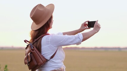 A woman in a hat stands in a field, capturing a beautiful sunset with her smartphone. The scene conveys calmness and nature
