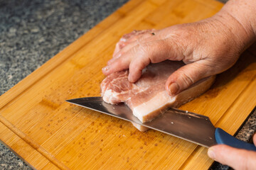 An adult hand is shown cutting a piece of raw pork with a knife on a wooden cutting board in a domestic kitchen setting.