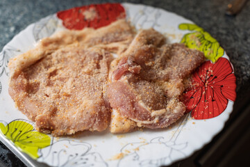 Raw meat cutlets coated in breadcrumbs are arranged on a white plate with colorful floral patterns, ready for cooking.