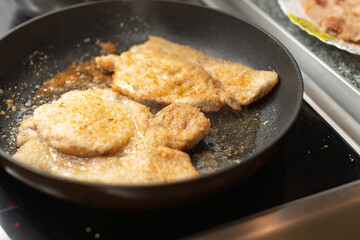 Several breaded cutlets are frying in hot oil in a black pan on a stovetop, turning golden brown as they cook.