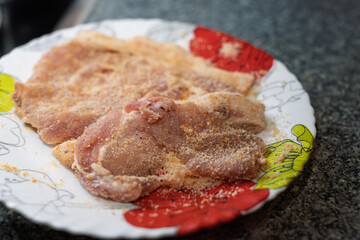 Several raw meat cutlets, coated in breadcrumbs and spices, are arranged on a decorative plate, ready for cooking.