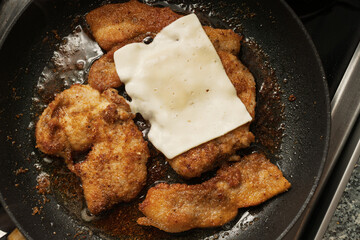 Golden-brown breaded meat pieces are frying in a dark pan, with one piece topped with a slice of melting white cheese.