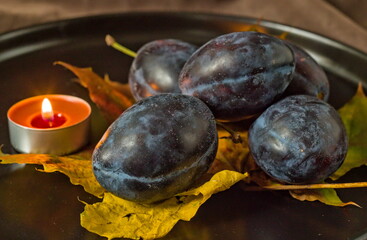 A warm and romantic autumn still life. Ripe purple plums are arranged with a yellow fallen leaf, while the soft, flickering glow of a lit tea light candle creates a cozy and intimate  atmosphere.