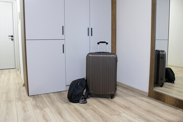 A bright apartment hallway: a rolling suitcase and backpack beside a wardrobe and tall mirror. Theme of arrival check out, short-term rental, business or leisure visit. © Elena