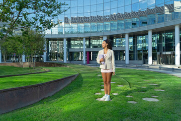 Young woman pausing outdoors, holding a folder, standing on a green lawn near a contemporary glass...