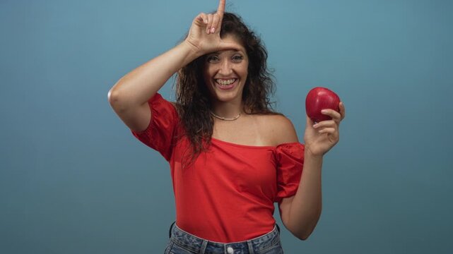 Woman holding a red apple and making an l sign on her forehead, bare shoulder visible in blue studio; playful teasing.