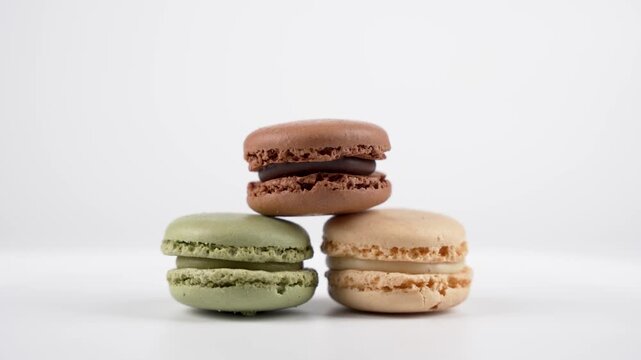A single chocolate, pistachio, and vanilla macaron stack rotates slowly on a clean white background. The smooth movement highlights texture and color.