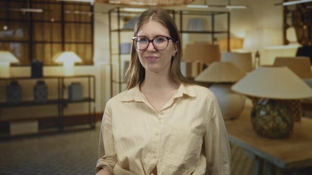 Young blonde woman wearing glasses and a beige shirt shows middle finger beside modern lamp display in a spacious building; defiance anger rejection contempt.