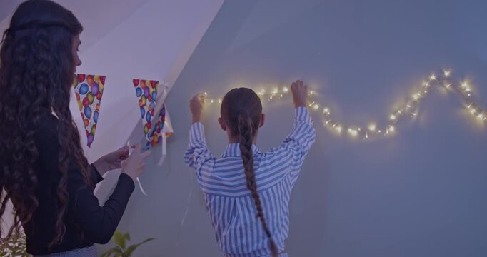 Young girls happily decorating a cozy room with glowing string lights and colorful banners. Preparing for a fun birthday party celebration at home, creating a warm and festive atmosphere.