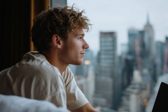 A man in a white shirt is looking out the window of a hotel room at a city - Powered by Adobe
