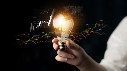 A person holds a glowing light bulb symbolizing innovation in a dark setting filled with financial...