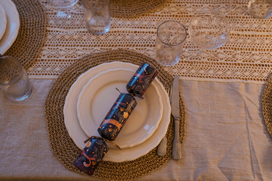 Elegant Christmas table setting with white plates, woven placemats, crystal glasses and festive crackers on a linen tablecloth with crochet details.